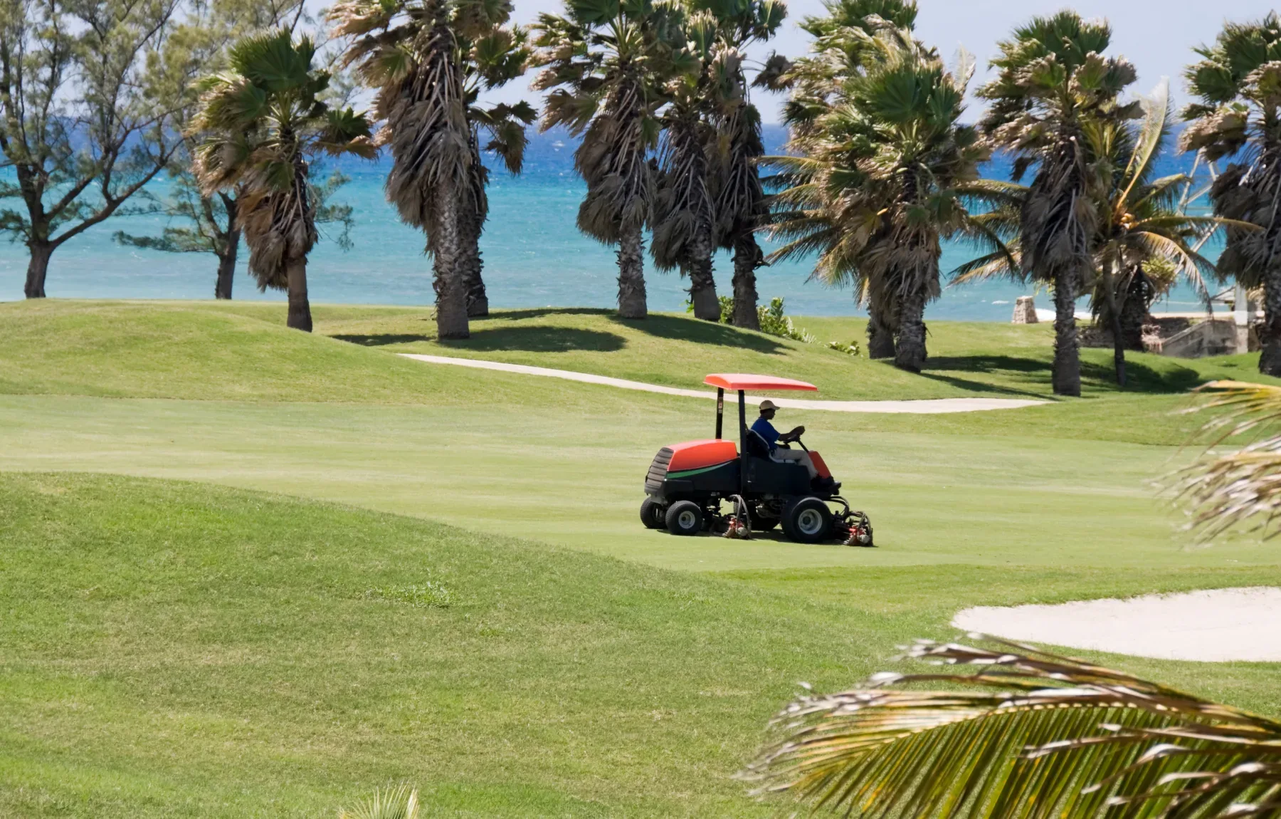 A groundskeeper drives a reel mower with a red sun canopy across a golf course fairway, cutting turf near the green. Rows of