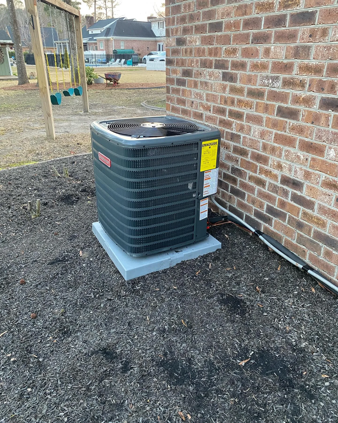 New HVAC air conditioning unit installed against brick home exterior with yard in background.