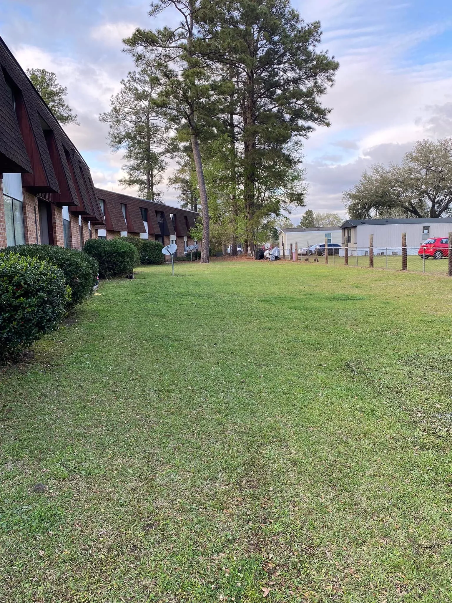Large open lawn with brick building and tall pine trees in background.