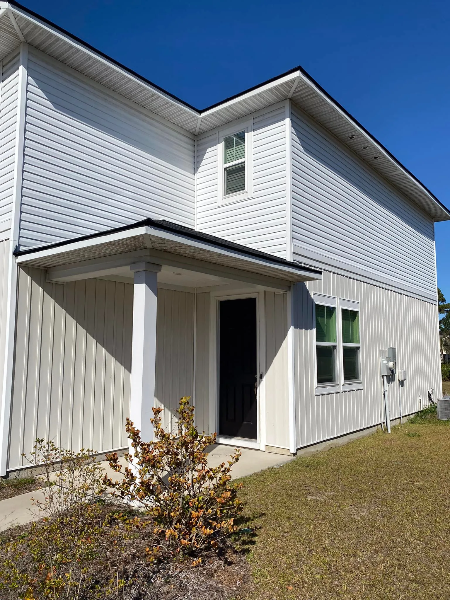 Modern white metal-sided house with covered porch and landscaped yard under clear blue sky.